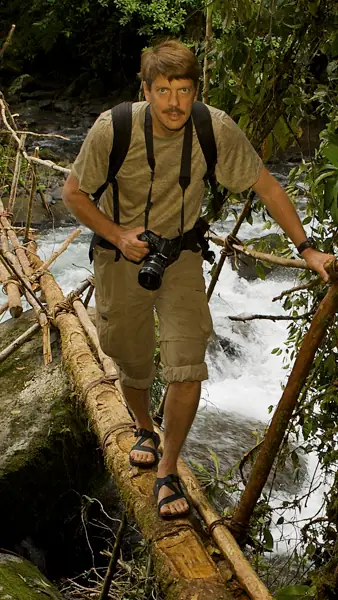 Tim Laman crossing a log bridge over a rainforest stream with a camera in his hand