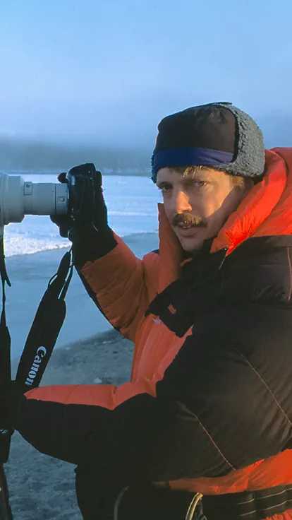 Tim Laman in a red down parka behind a long telephoto lens on a tripod, beside a frozen lake